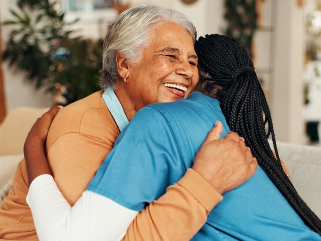 Two women share a warm hug, smiling in a bright, cozy room, suggesting care and connection.