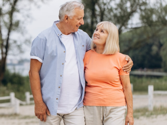 Older couple walking together outdoors, smiling and looking at each other, with arm around each other in a park setting.